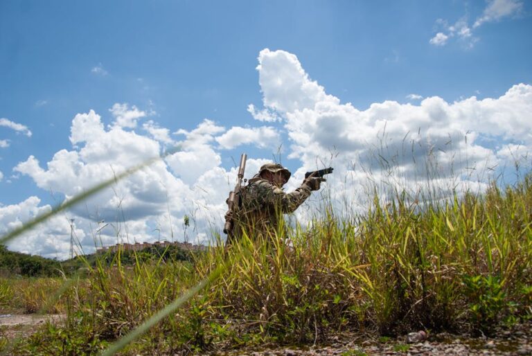 Soldier in camouflage navigating grassy field under clear blue sky, perfect for tactical and military themes.