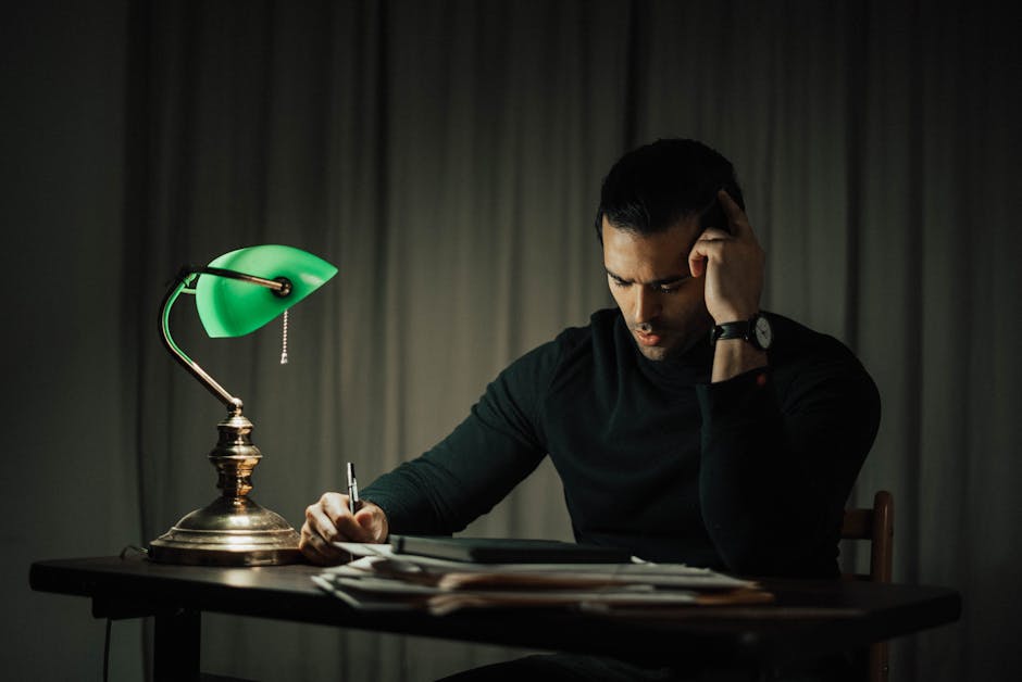 Focused man working late at a desk, lit by a characteristic green banker’s lamp, surrounded by paperwork.