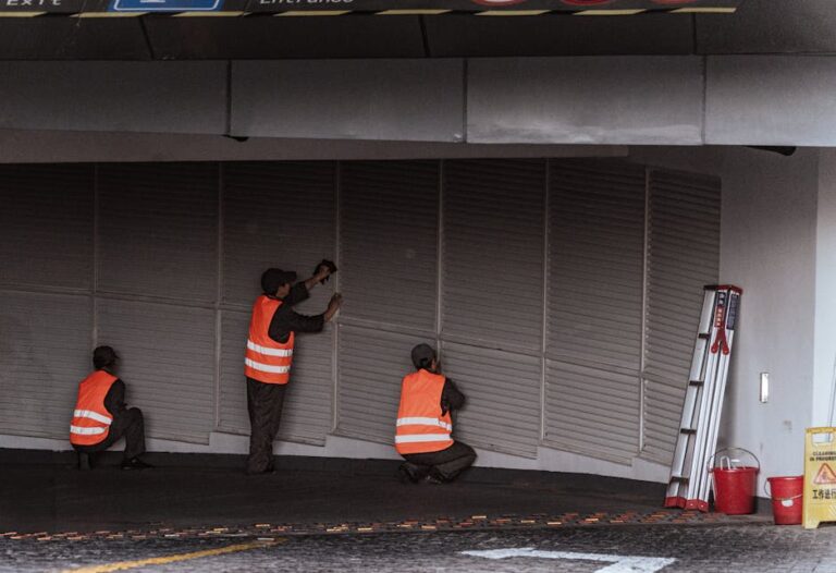 Three workers in safety vests install security shutters at an indoor entrance.