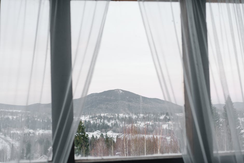 A serene winter landscape framed by sheer curtains, showcasing snow-covered mountains and forests.