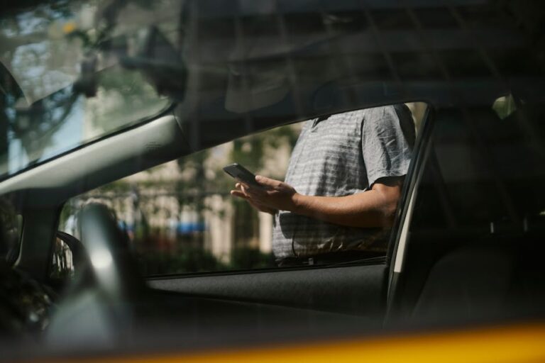 A man using a smartphone inside a car, visible through the window in an urban setting.