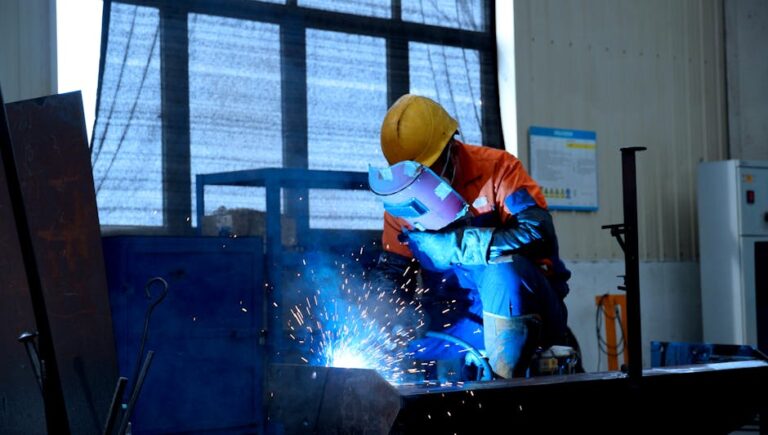 A welder in protective gear works in a factory, creating bright blue sparks.