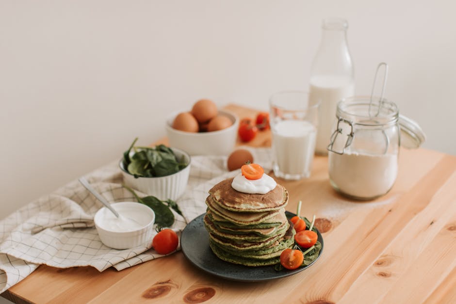 Delicious stack of pancakes with fresh spinach, tomatoes, eggs, milk, and yogurt on a wooden table.