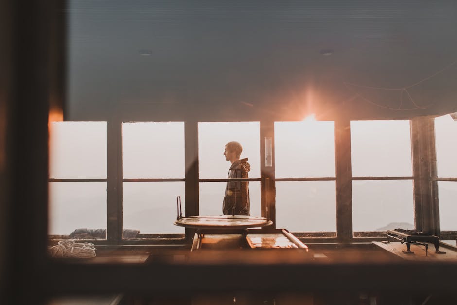 A contemplative man stands in a sunlit room with large glass windows, capturing a serene moment.