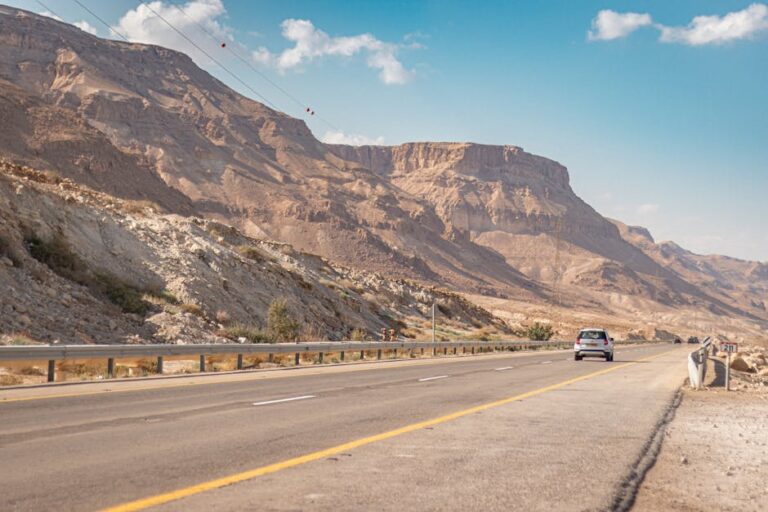A car travels on a scenic road through desert mountains under a bright blue sky.