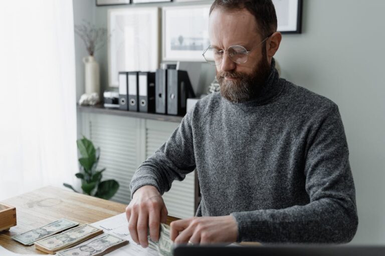 An adult man in a gray sweater counting dollar bills at a wooden desk, showcasing financial management.
